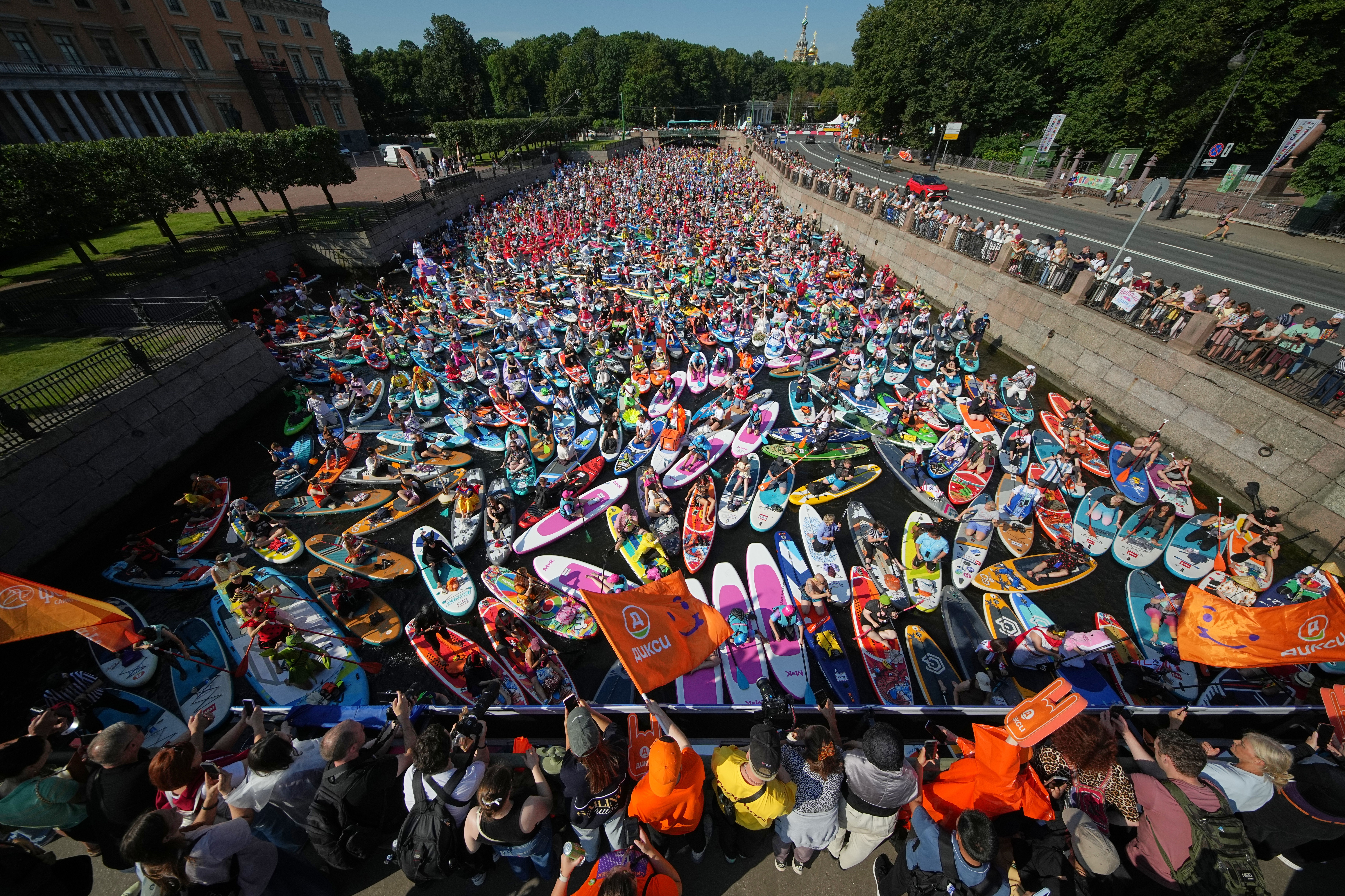 Hundreds of people sit and stand on paddle boards, crowded together on a river in St. Petersburg, Russia.