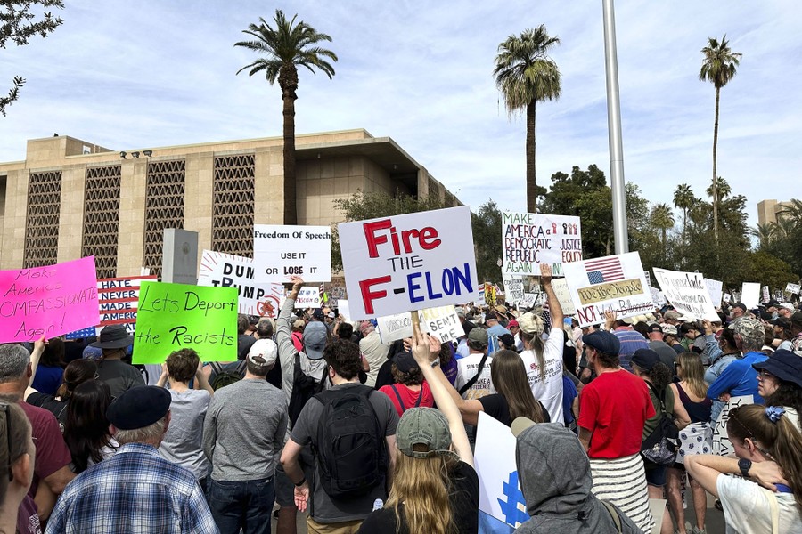 A group of protesters gather outside a capitol building, holding signs, including one that reads 