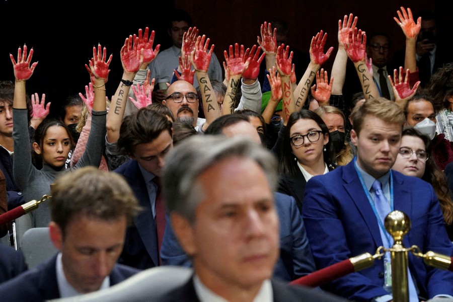 Protesters raise their hands, palms covered in red paint, while sitting and watching a committee meeting.
