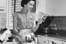 A woman reads a cookbook next to a mixer.