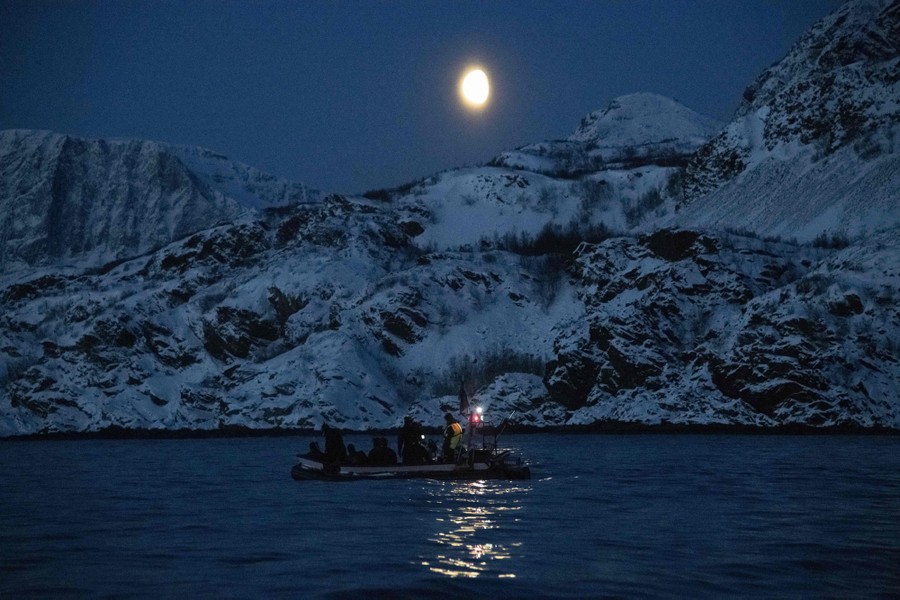 Several people float in an inflatable boat on a fjord at night, with snow-covered mountains in the background.