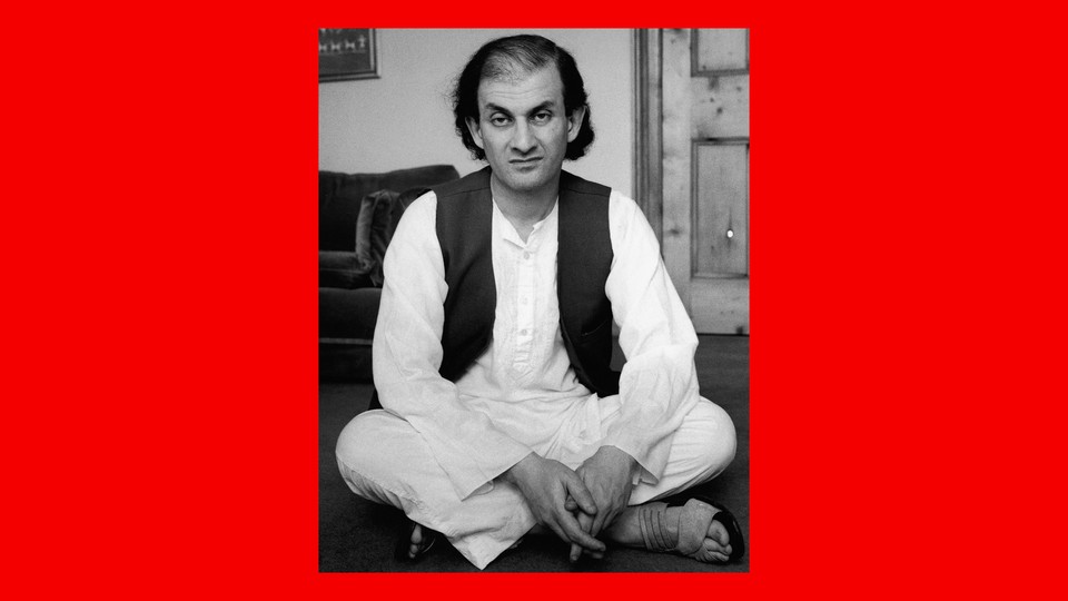 In a black-and-white image from 1988, Salman Rushdie, with chin-length dark hair and wearing a dark vest over white shirt and pants, sits crossed-legged on the floor.