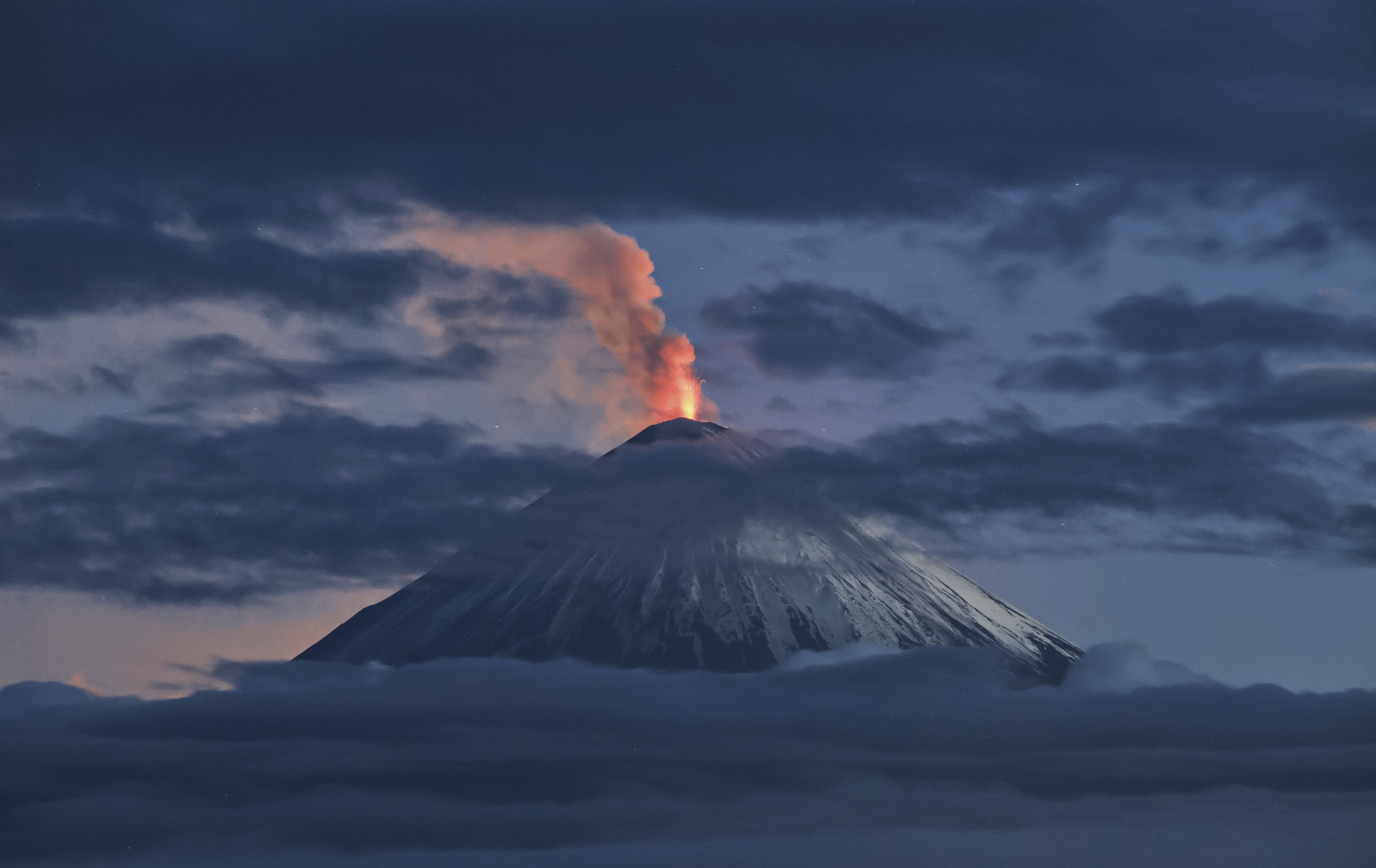 A distant view of an erupting cone-shaped volcano, among clouds