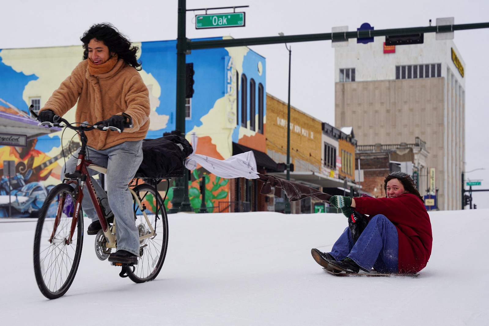 A person rides a bike, pulling a person behind them over a light dusting of snow.