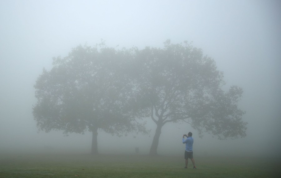 A man takes a photograph in a park amid thick fog.