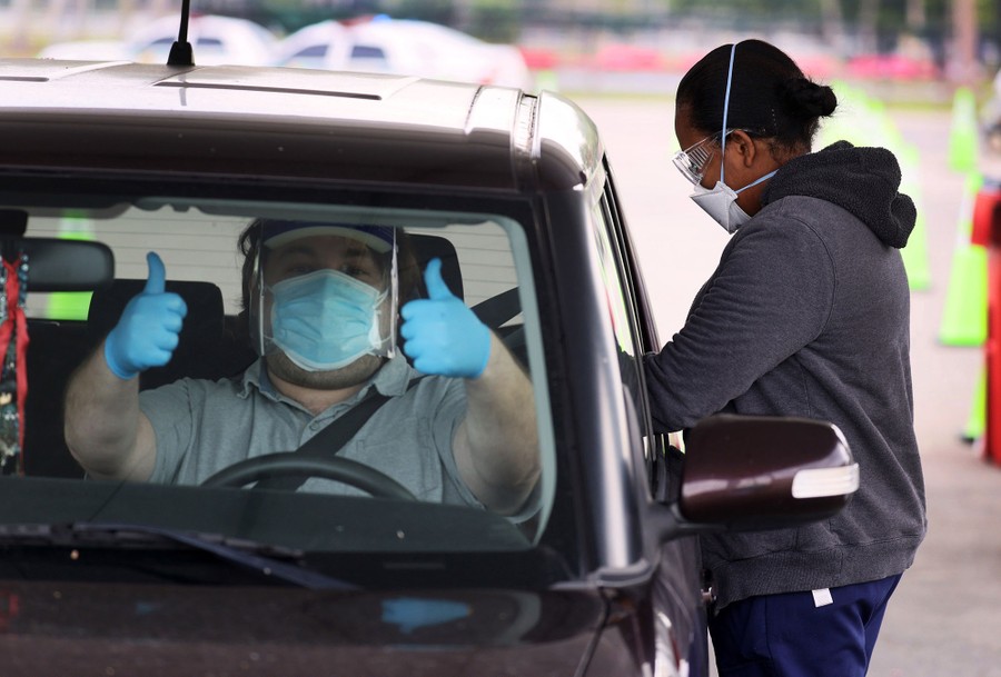 A masked man in the driver's seat of a car gives a thumbs-up to the camera as a worker gives a dose of vaccine to a passenger in the car's back seat.
