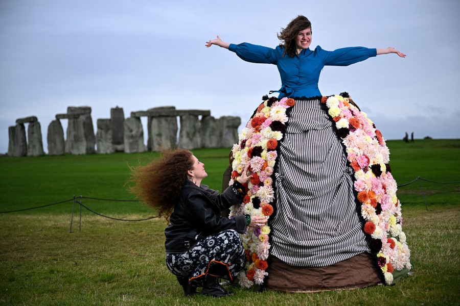 One person stands in a dresslike structure, partly covered in dahlias, while another person adjusts the dress, in front of Stonehenge.