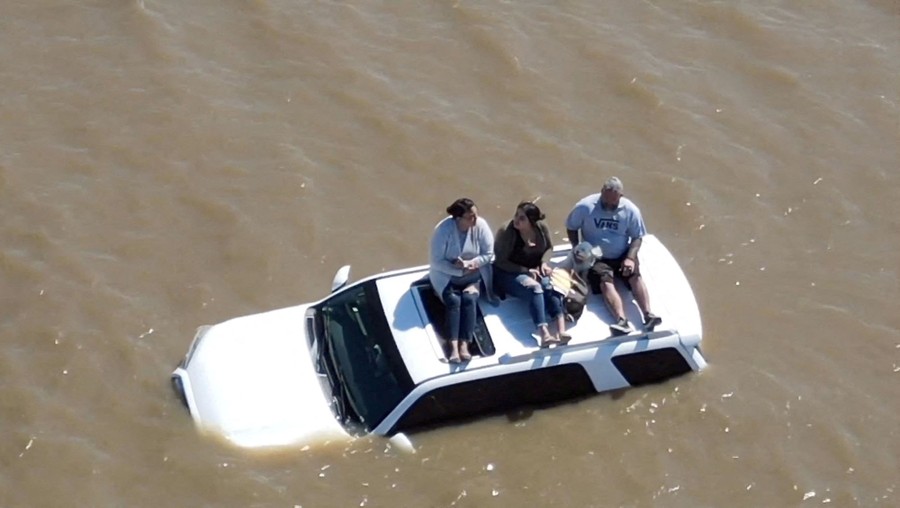 Three people sit on the roof a car that is partially submerged in floodwater.