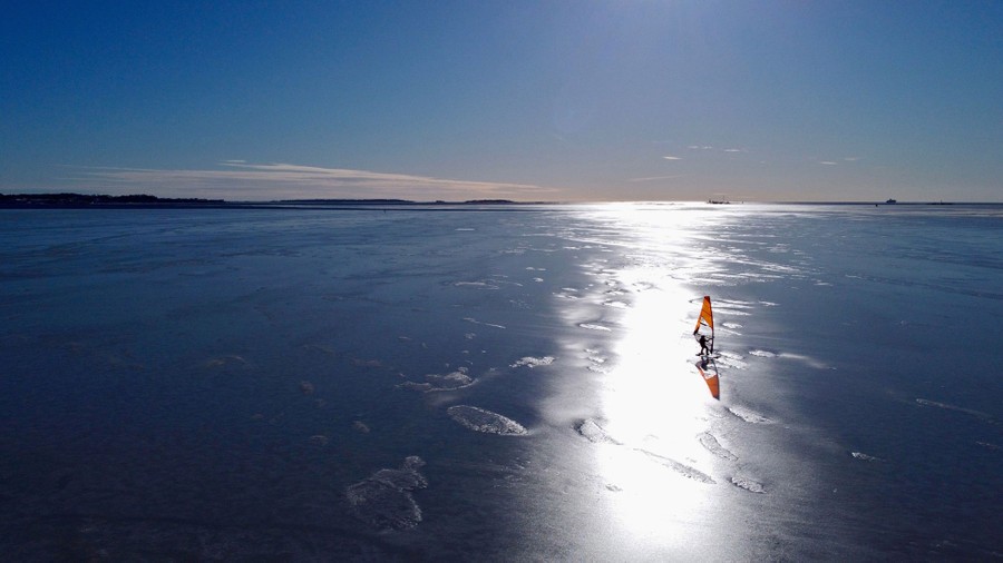 A person skates across a vast sheet of ice, holding a sail that is pushed by the wind.