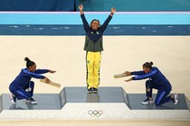 On a medals podium, two gymnasts on either side of the gold-medal winner playfully bow down to her, as she raises her arms.