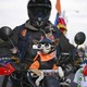 A motorcyclist rides in a caravan with a small dog resting on their handlebars. The dog wears a leather vest, a bandana, a helmet, and doggles.