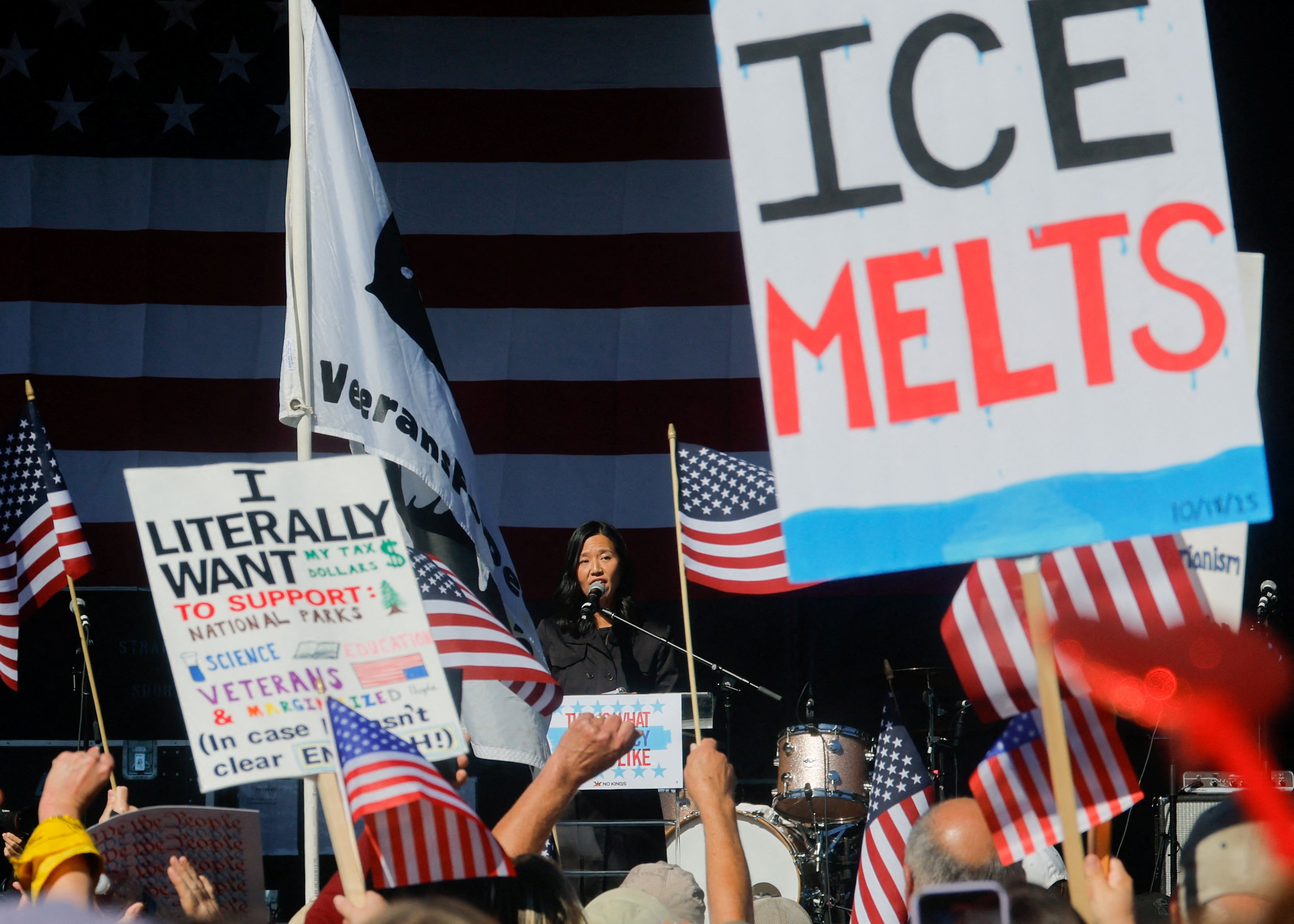 A crowd of protesters watches the Mayor of Boston speak at a podium, holding signs, including one that reads