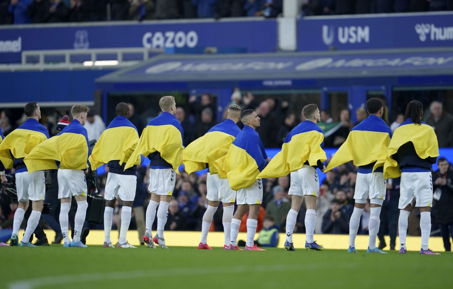 Soccer players wearing Ukrainian flags draped over their shoulders line up on a pitch.