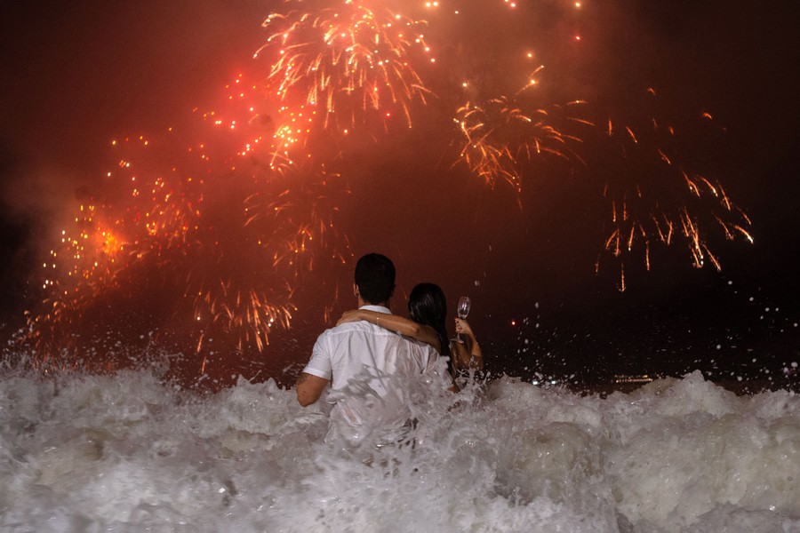 Two people stand in crashing surf while watching fireworks.