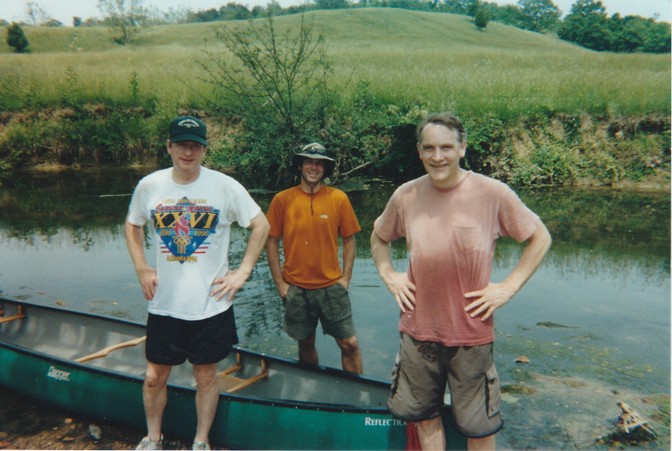 Three men standing next to a canoe by a river with a hilly landscape in the background