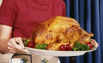 A person in a pink shirt and white skirt holding a tray with a roasted turkey on it