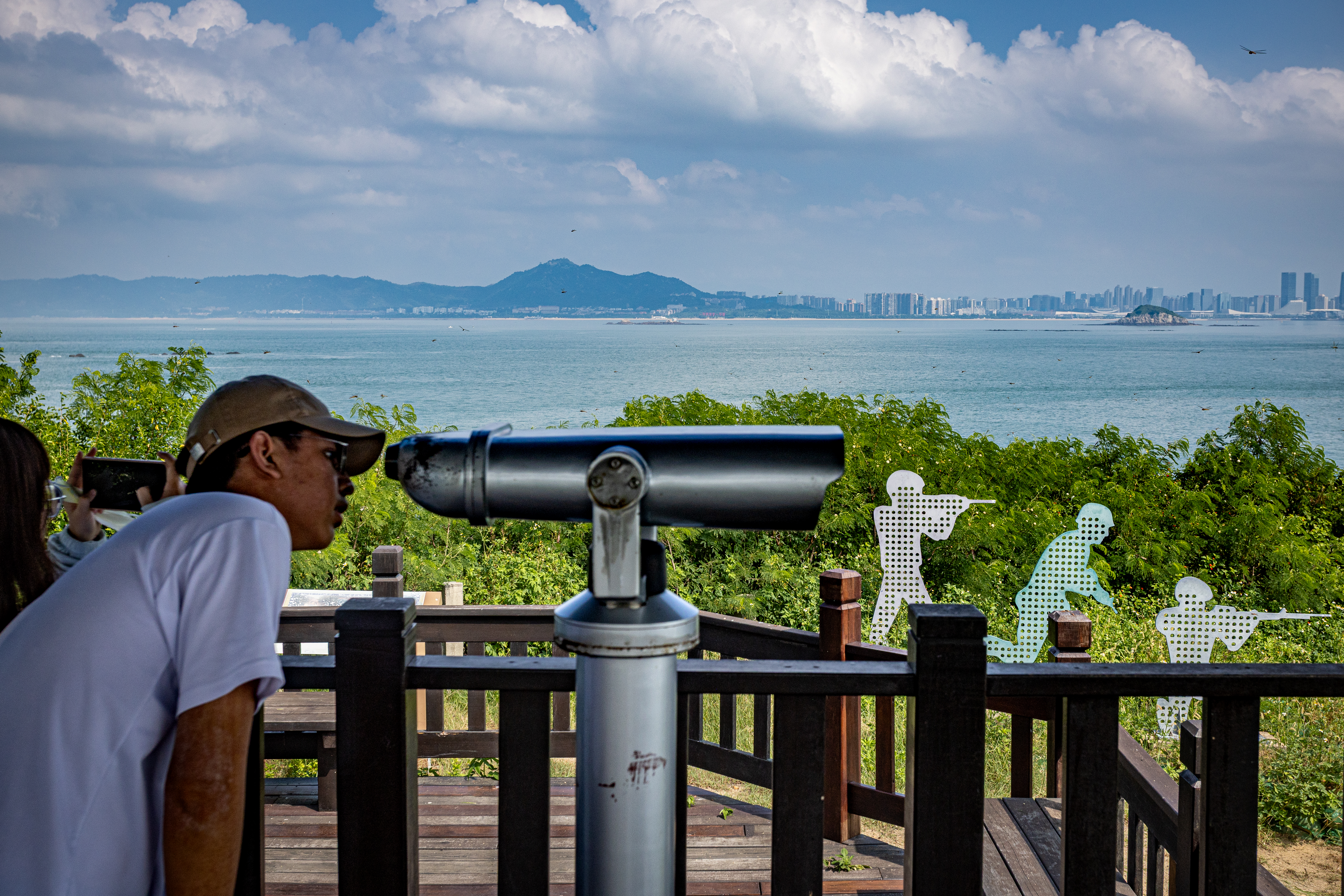 male tourist looks through telescope across view of shore, water, city skyline, and mountains in distance, with several silhouette sculptures of military figures aiming and firing weapons in foreground