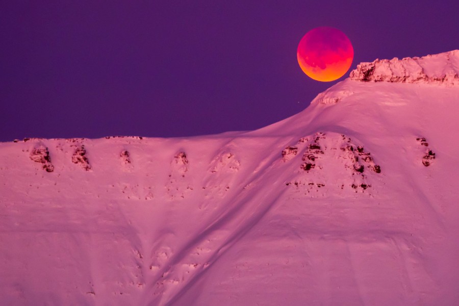 A partially obscured, reddish moon over a large, snow-covered mountain