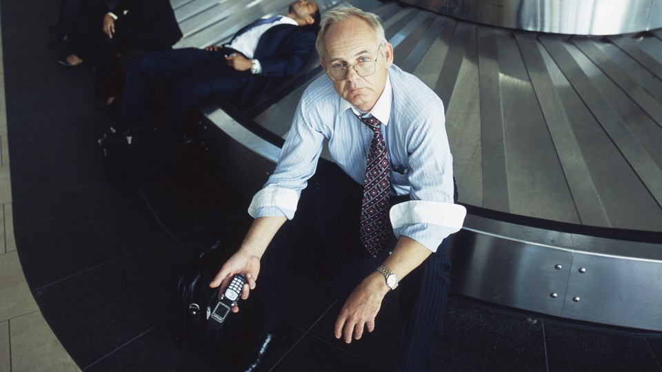 A man in a shirt and tie holds a cellphone and looks upset while sitting on a baggage carousel at an airport.