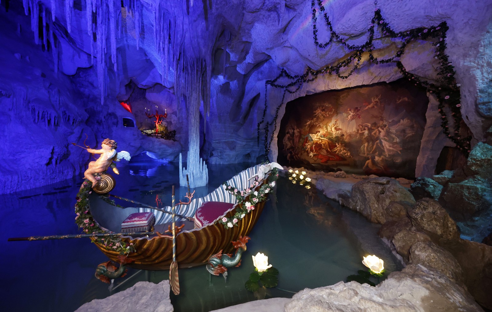 A small ornate and fanciful boat sits in water inside an artificial grotto, theatrically illuminated.