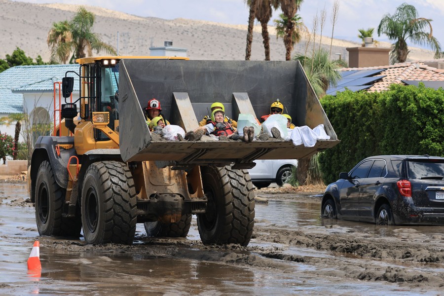 Three rescue workers sit in the bucket of a front-end loader, each of them holding a civilian in their lap, as they are carried out of a flooded area.