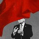 Photo of a man in a suit clapping with a huge red flag in the foreground, covering half his face.