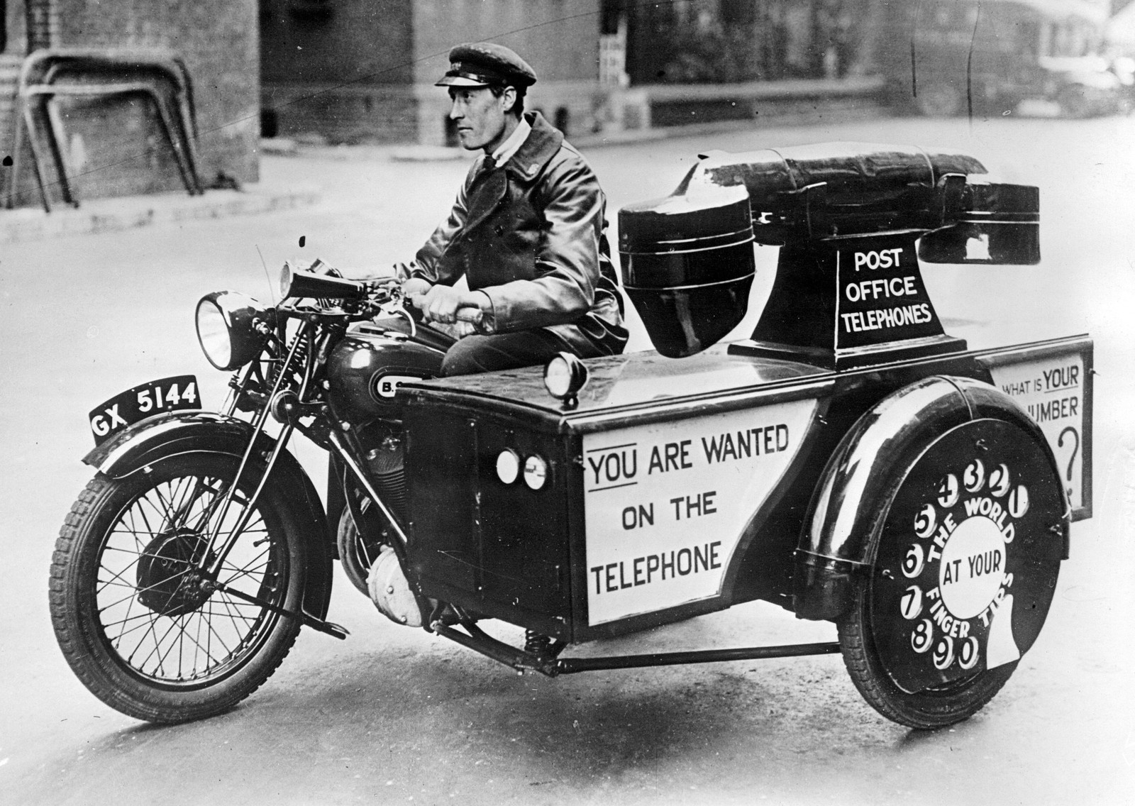 A man drives a motorcycle with a side car that is shaped like a gigantic telephone, with the phrase