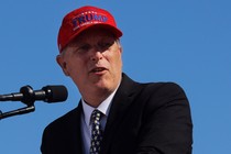 Andy Biggs stands before a microphone wearing a "Trump: Make America Great Again" hat.