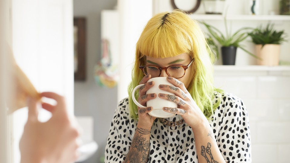 A young woman with dyed hair and tattoos sips a cup of coffee.
