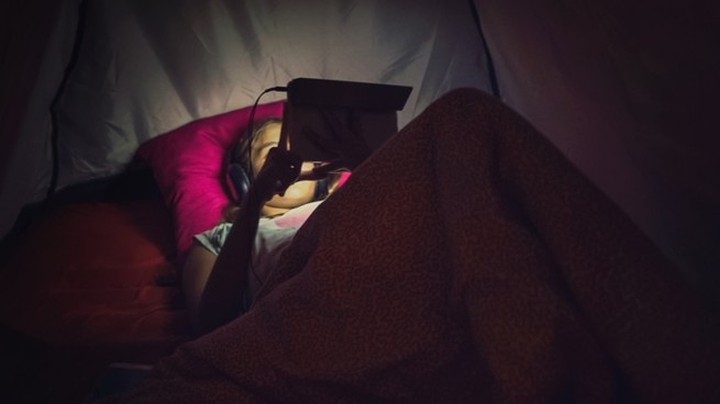 A child lying in bed, playing on their iPad.