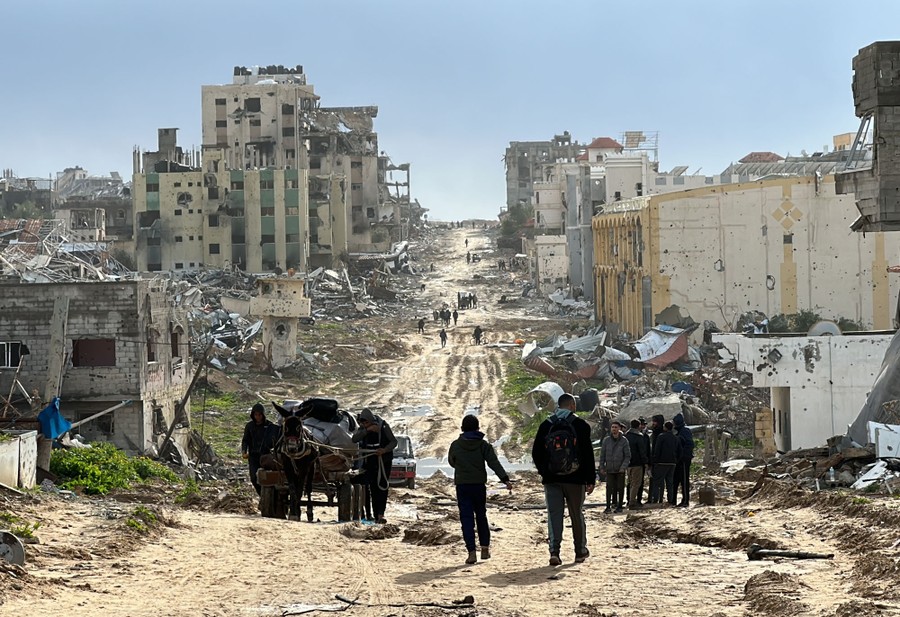 People walk on a dirt road past many damaged and destroyed buildings.