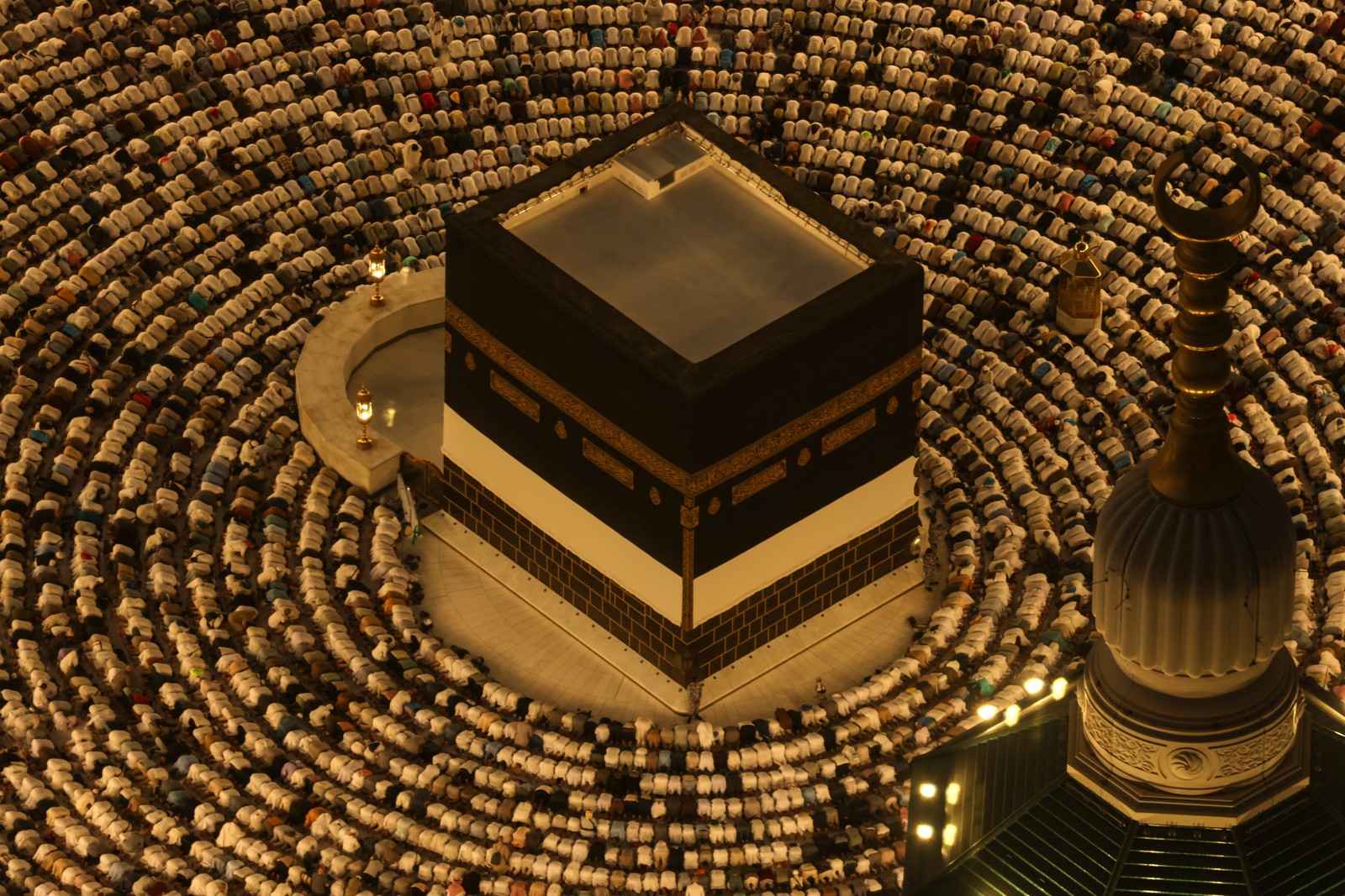 An elevated view of hundreds of people kneeling in prayer in concentric circles around the Kaaba, a large cube-shaped structure.