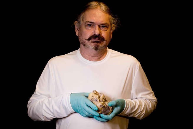 A nan wearing a white shirt and blue latex gloves holds his own heart in front of his chest.