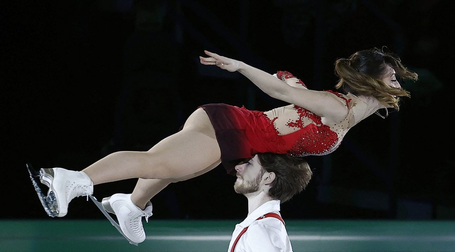 A figure-skating couple perform. One is held horizontally aloft by the head of her partner, pressed against her belly.