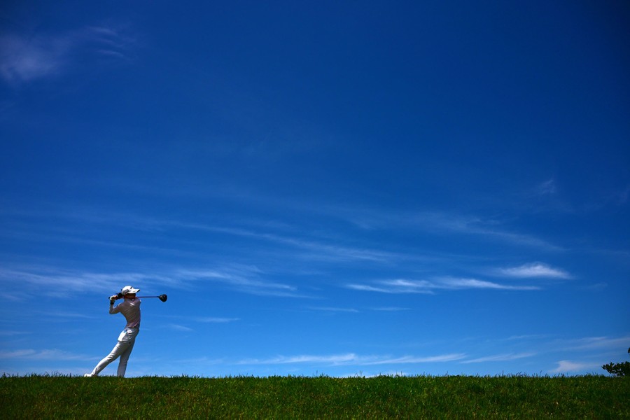 A golfer hits a shot, seen against a large expanse of blue sky.