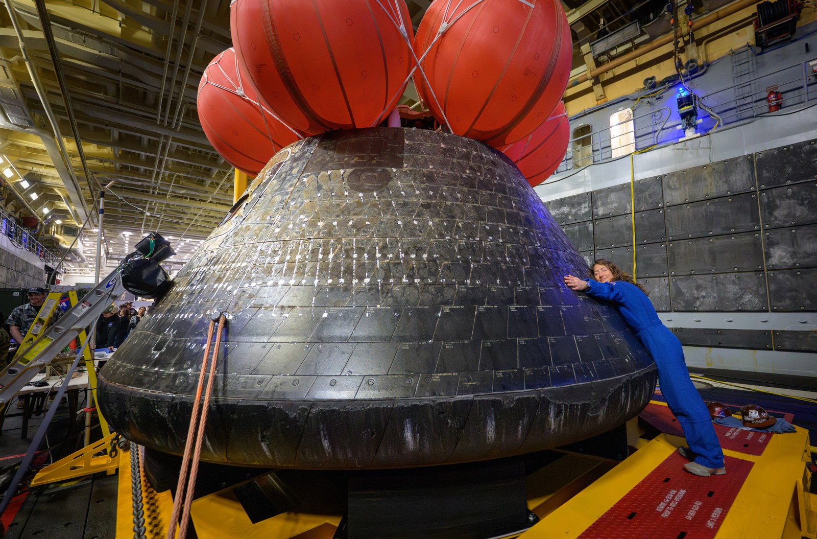 An astronaut, wearing a blue jumpsuit, leans on and embraces the space capsule she returned to earth in earlier.