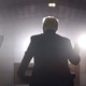 Donald Trump's back as he stands at a lectern, lit from the front
