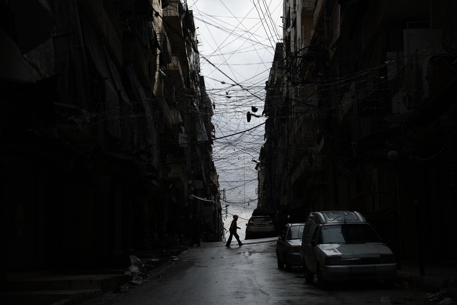 A man crosses a street, flanked by tall, dark buildings and a chaotic web of wires above.