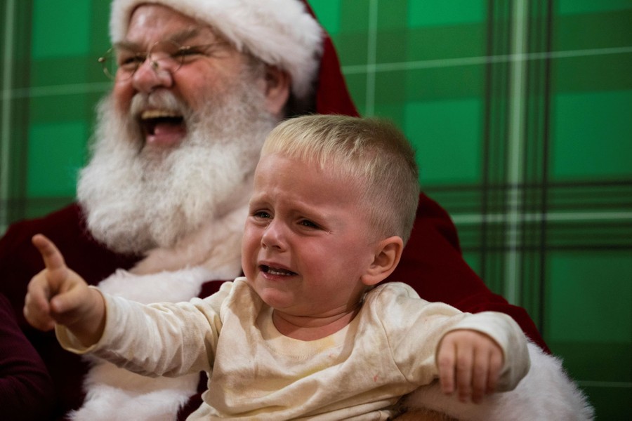 A young child cries while sitting on Santa's lap. Santa laughs.