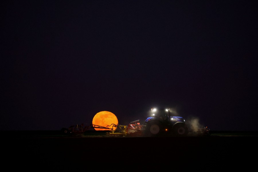 A farmer drives a tractor in a field, with the full moon on the horizon.