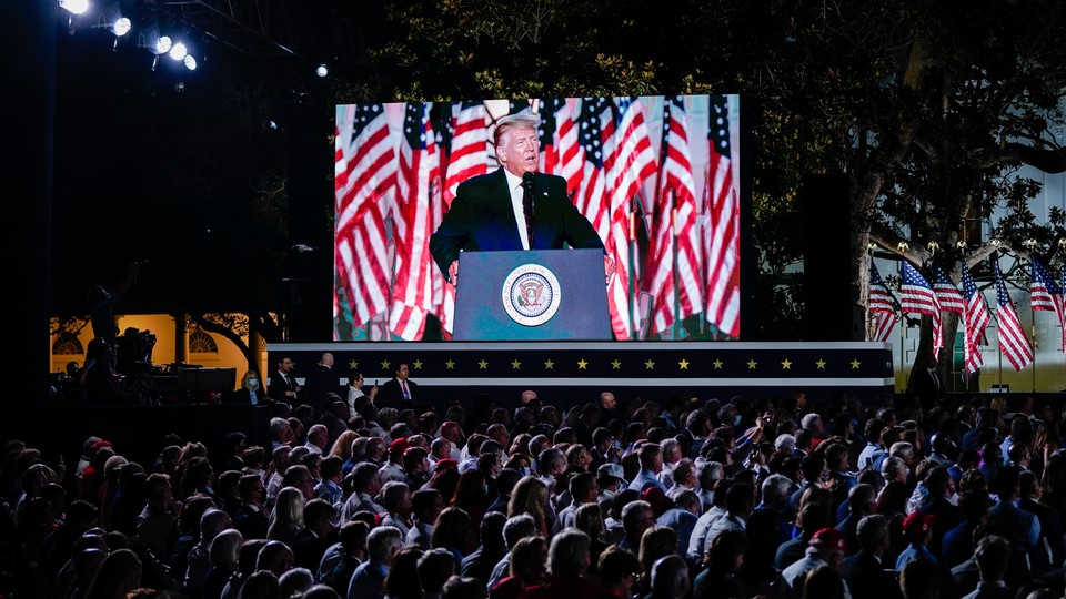 A crowd stares up at President Donald Trump while he makes a speech behind a podium. Trump is visible on a giant screen to the crowd's left.