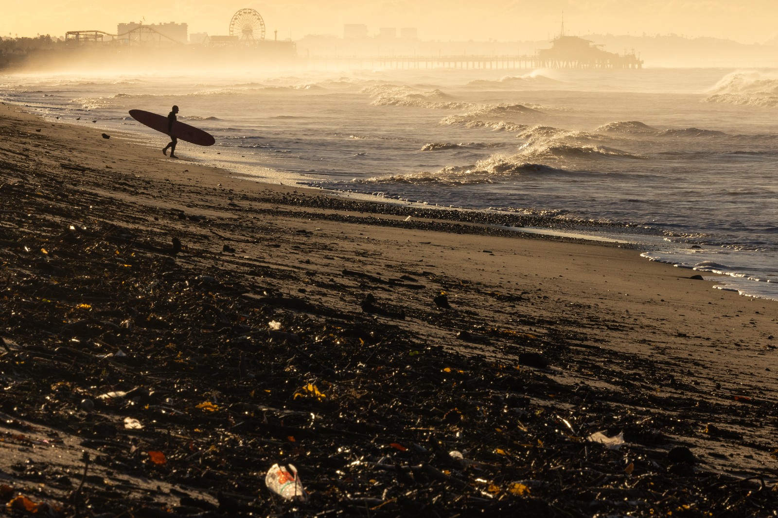 A person carries a surfboard toward the shoreline, walking on a beach that is partly covered with debris from a previous wildfire