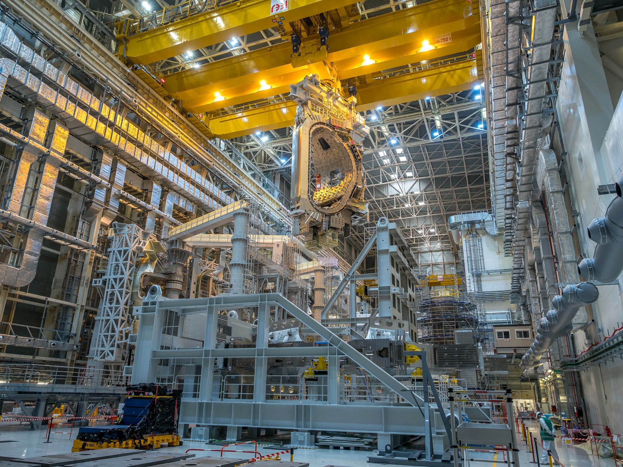 A large D-shaped component of a fusion reactor is lowered into place among many supports and other partially complete components.