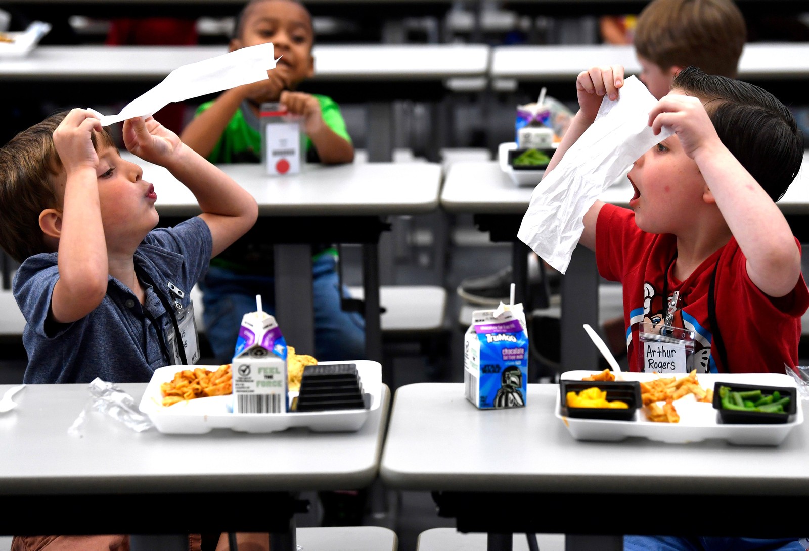 Two boys sitting at cafeteria tables play with each other, blowing on napkins.