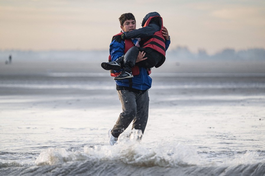 A person carries a child wearing a flotation vest through shallow surf.