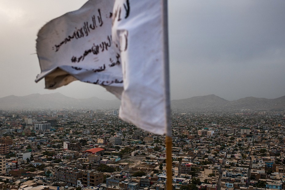 A frayed white flag with black Arabic writing flies over a dense city landscape, with distant hills in the background