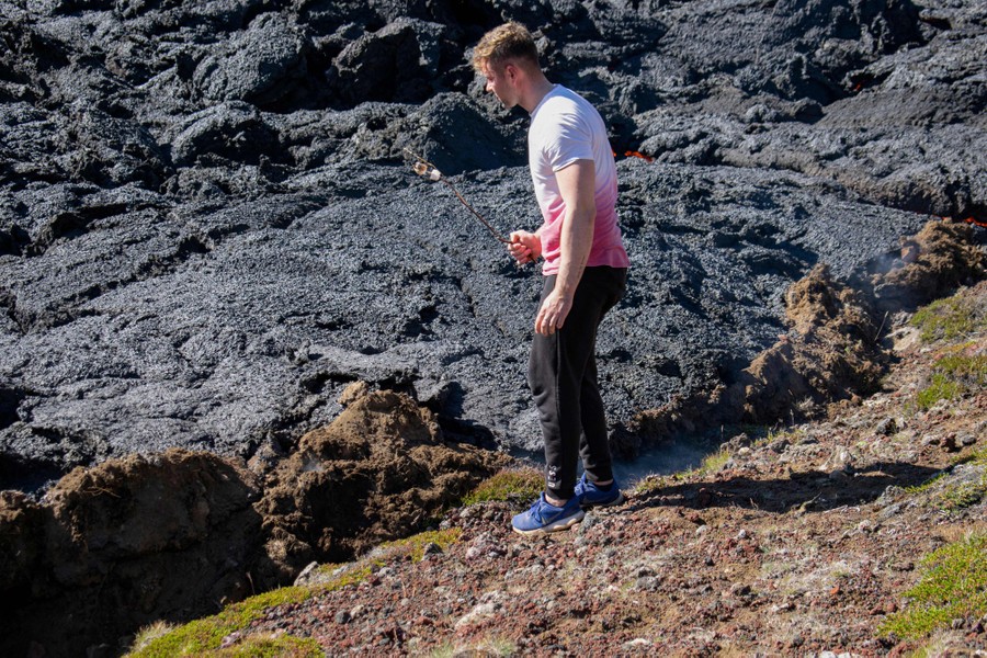 A person stands very near a lava flow holding a stick in hand with two marshmallows on the end.