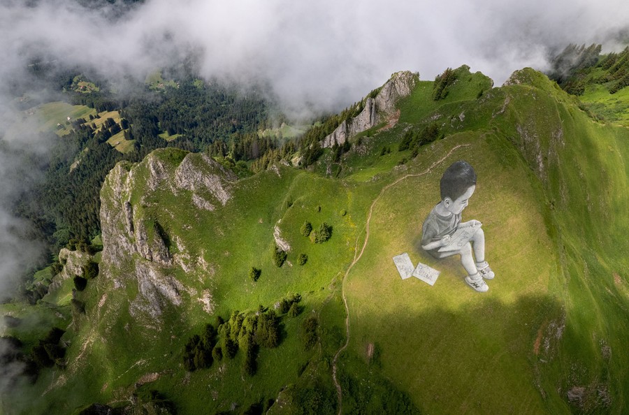 An elevated view of a huge mural of a seated child that has been painted onto the grassy slope of a mountain