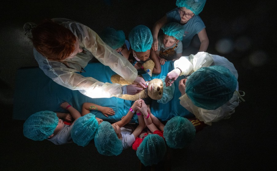 A top-down view of two adults and nine young children wearing protective medical gear, all leaning forward to attend to a teddy bear lying on a gurney.