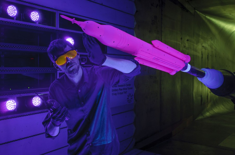 A person wearing goggles works alongside a four-foot-long scale model of a rocket. The small room is lit by black light, and the rocket glows pink.